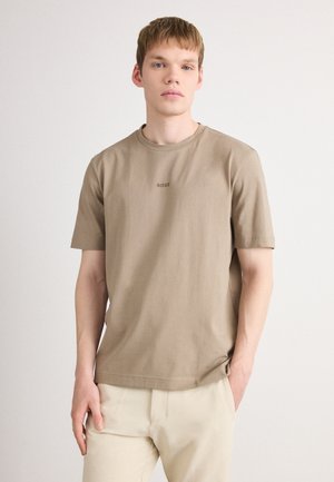 Junger Mann mit hellen Haaren, der ein beiges BOSS-T-Shirt und cremefarbene Hosen trägt, steht mit einer Hand in der Tasche vor einem schlichten Hintergrund.