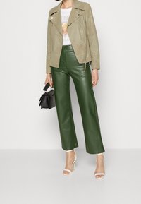 Pantalons larges en simili cuir vert olive associés à une veste de motard en suède beige clair, un T-shirt graphique blanc et des sandales à talons blanches.