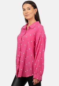 Camicia oversize rosa con bottoni in tessuto testurizzato e motivo a cuori bianchi, maniche lunghe e colletto classico. Indossati sotto, leggings neri.