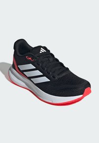 Chaussure de sport noire avec tige en maille, rayures blanches, accents rouges, semelle rembourrée et tissu texturé. Présente des lacets et un logo sur la languette.