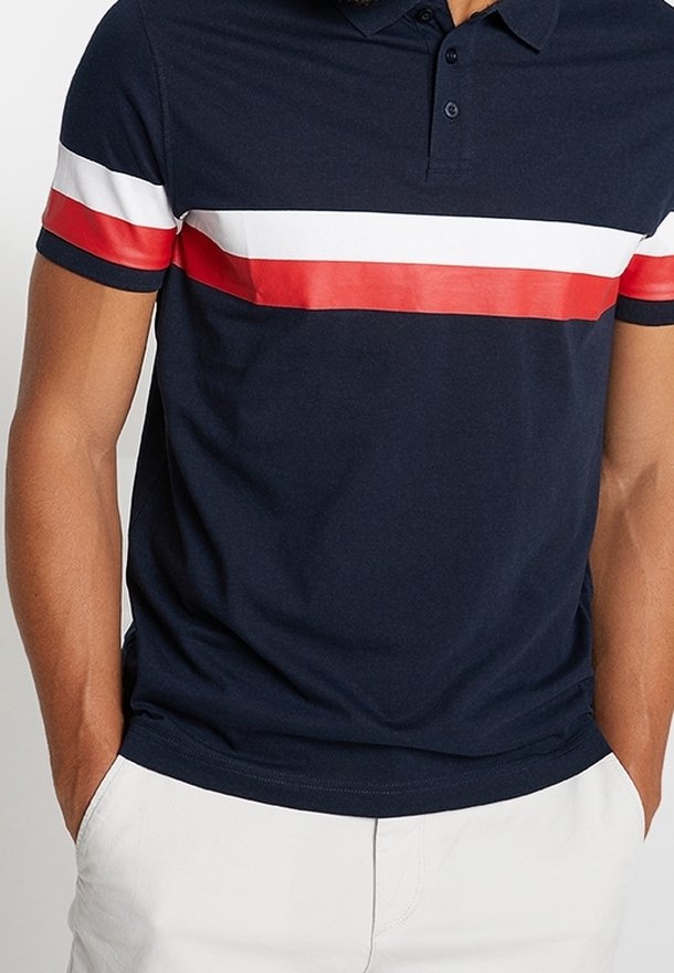 Homme portant un polo bleu marine avec des rayures horizontales blanches et rouges sur la poitrine et les manches, associé à un pantalon blanc.