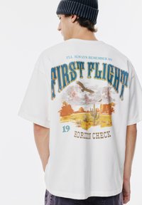 Weißes, übergroßes T-Shirt mit einem bunten "First Flight"-Grafikdesign, Wüstenlandschaft mit Kakteen, Adler und Textakzenten in Blau und Orange.