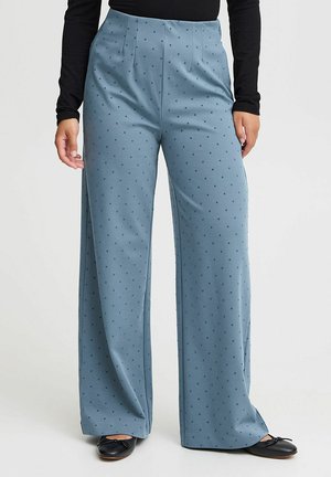 Wijdvallende broek in lichtblauwe stof met donkerdere stippen, met een hoge taille en plooidetails, gecombineerd met zwarte ballerina's.
