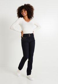 Top blanco de manga larga con bordes de encaje, combinado con unos vaqueros oscuros de tiro alto que tienen dos botones dorados y costuras decorativas. Zapatillas blancas.