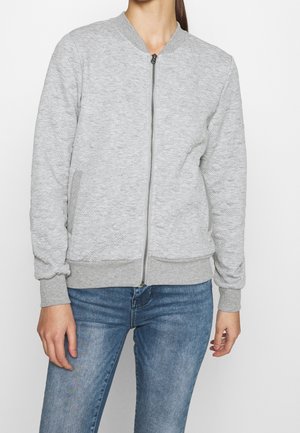 Mujer que lleva una chaqueta bomber con cremallera en gris claro texturizada, con puños y dobladillo de canalé, combinada con vaqueros azules.