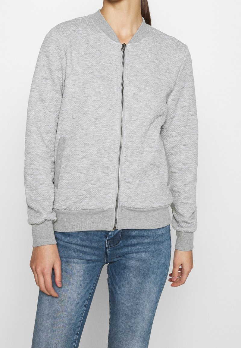 Femme portant une veste bomber zippée gris clair texturée, avec poignets et ourlet côtelés, associée à un jean en denim bleu.