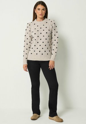Frau trägt einen beigen Pullover mit schwarzen Punkten, schwarze Hose und beigefarbene Schuhe, steht aufrecht vor einem einfachen hellen Hintergrund.