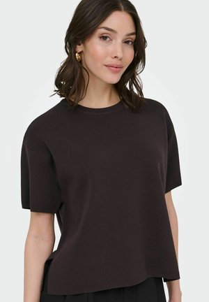 Jeune femme aux cheveux bruns mi-longs portant des boucles d'oreilles en or en forme de créole et un haut noir à manches courtes et amples, se tenant devant un fond clair.