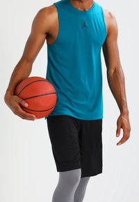 Uomo che indossa una maglietta senza maniche color turchese e pantaloni corti neri, mentre tiene un pallone da basket su uno sfondo chiaro e semplice.