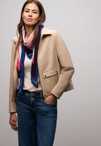 Beige uldblandet jakke med krave og sidelommer, stylet med et farverigt, mønstret silketørklæde og blå denimjeans.