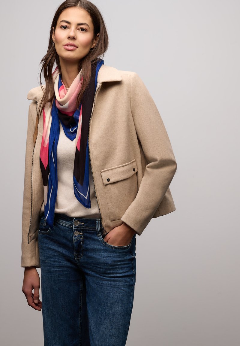 Beige uldblandet jakke med krave og sidelommer, stylet med et farverigt, mønstret silketørklæde og blå denimjeans.