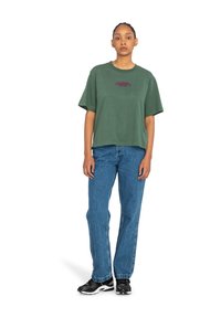 T-shirt verde oversize con maniche corte e scollatura rotonda, con testo color marrone scuro. Abbinato a jeans dritti azzurri chiari e sneakers nere.