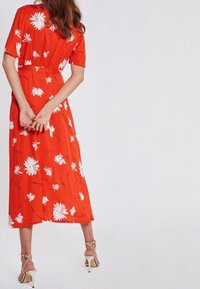 Robe midi rouge à fleurs avec manches courtes, motif marguerites blanches, taille cintrée et silhouette fluide, assortie à des talons clairs.