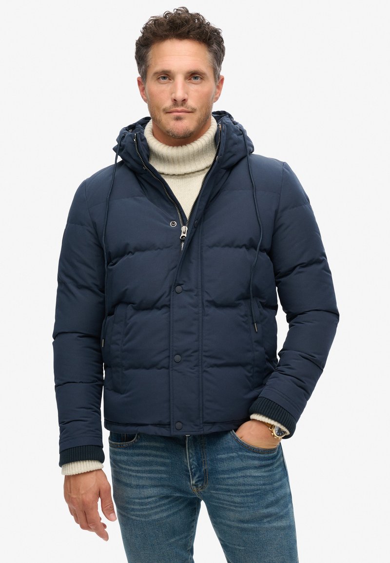 Chaqueta de abrigo en color navy con capucha, cierre de cremallera y botones a presión. Puños y dobladillo acanalados, llevada sobre un suéter de cuello alto color crema.
