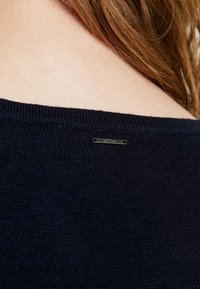 Granatowy sweter dzianinowy z szerokim dekoltem i małą, srebrną metką marki z tyłu. Gładka faktura i minimalistyczny design.