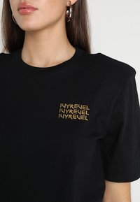 Černé bavlněné tričko s vyšívaným zlatým logem na levé části hrudi. Klasický kulatý výstřih a krátké rukávy. Měkká textura.