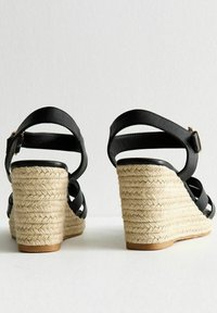 Black leather wedge sandals with woven jute heel, featuring crisscross straps and a buckle closure at the ankle.