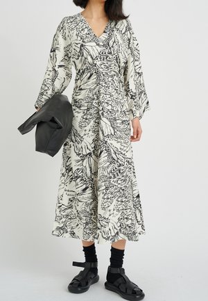 Femme portant une longue robe à imprimé abstrait noir et blanc, des chaussettes noires, des sandales noires, et tenant une veste en cuir noire pliée.