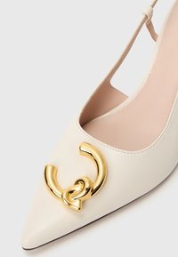 Valkoinen nahkainen kärkikolmiokenkä, jossa on kullanvärinen metallikoriste, jossa on toisiinsa kytkeytyviä pyöreitä muotoja ja kermaisen sileä pinta.