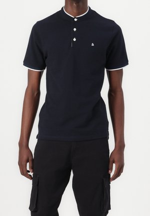 Homme portant une chemise bleu marine à manches courtes avec un liseré blanc au col et aux manches, trois boutons, et un pantalon cargo noir avec des poches.