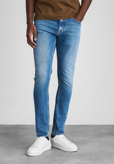 Vaqueros de mezclilla azul claro con corte recto, que presentan áreas deslavadas y diseño clásico de cinco bolsillos, combinados con zapatillas blancas.