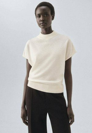 Pull en maille crème avec un col haut et des manches courtes, associé à un pantalon sombre. La texture semble douce et lisse.