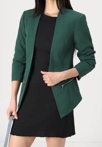 Zelený blazer s přiléhavým střihem, dlouhými rukávy a stříbrnými zipovými detaily na bocích, nošený přes černé šaty. Hladká textilie.