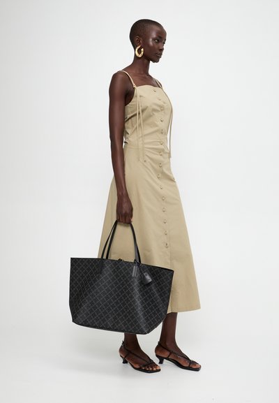 Black patterned tote bag with a structured shape, leather handles, and a charm. Worn with a beige buttoned dress and strappy black sandals.