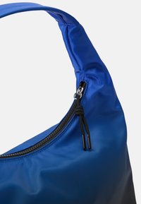 A mala de ombro azul em nylon apresenta uma forma curva, uma alça plana e um fecho zip preto com puxador em corda. Uma textura suave domina a superfície.