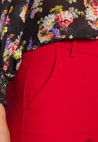 Blouse florale avec fond noir et fleurs colorées, associée à un pantalon rouge ajusté avec une poche latérale et une texture lisse.