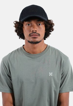 Homme aux cheveux bouclés portant une casquette noire et un t-shirt vert olive à col rond avec un petit logo blanc sur la poitrine, face à l'avant sur un fond uni.