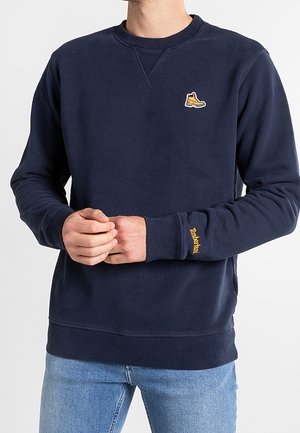 Mężczyzna ubrany w granatową bluzę z małym żółtym logo buta na piersi oraz napisem Timberland na mankiecie lewego rękawa, w połączeniu z jasnoniebieskimi dżinsami.