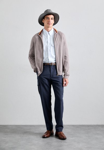 Cárdigan de punto gris claro, camisa azul de botones con bolsillo, pantalones cargo azul marino, cinturón con estampado y sombrero de fieltro gris; zapatos de slip-on marrones.