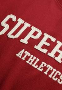 Sudadera roja con gran texto bordado en blanco que dice "SUPER" y "ATHLETICS" en una fuente audaz y bloque. Textura suave, diseño casual.