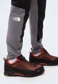 Grå og sorte North Face bukser kombineret med brune og sorte vandresko med orange accenter og Vibram-såler.