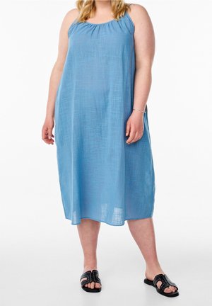 Kobieta ubrana w luźną, niebieską sukienkę midi bez rękawów oraz czarne wsuwane sandały z ćwiekami na białym tle.