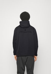 Mies kiharat hiukset, musta huppari ja tummat housut, seisoo selin yksinkertaisen vaalean taustan edessä.