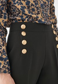 Schwarze Hosen mit hoher Taille verfügen über goldene Knöpfe an der Seite. Die Bluse hat ein floral gemustertes Design in Blau- und Brauntönen.