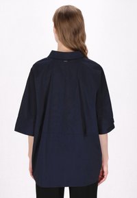Marineblaues Übergröße-Hemd mit Kragen, überschnittenen Schultern und kurzen Ärmeln. Glatte Textur mit einem Rückennahtdetail und dezentem Branding.