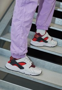 Sneakers hebben een witte bovenkant met rode en zwarte accenten, een gestructureerde zool en een opvallende hielcounter. Draagt het in combinatie met lavendelbroeken.