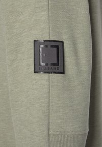 Lichtgroene trui met een gestructureerde stof en een klein zwart logo op de mouw, met een minimalistisch ontwerp.