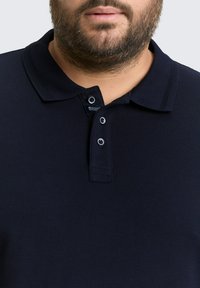 Marineblauw polo-shirt van een textuurstof, met een drieknoopsluiting en een geribde kraag. Bevat een label bij de halsopening.