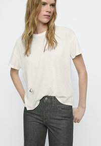 Lichtbeige t-shirt met korte mouwen, relaxte pasvorm, gecombineerd met donkergrijze jeans met zakken en gouden stiksels. Eenvoudige zilveren hanger.