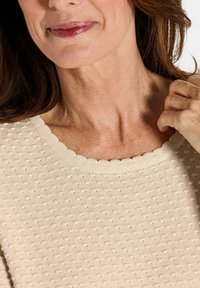 Close-up van de onderkant van het gezicht van een vrouw, haar nek en hand terwijl ze haar beige gestructureerde trui met een golvende halslijn aanpast.
