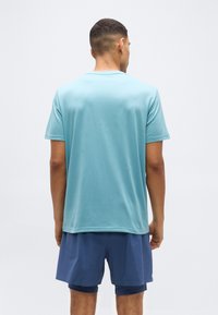 Άνδρας που φαίνεται από πίσω, φοράει ανοιχτό μπλε κοντομάνικο πουκάμισο και σκούρα μπλε αθλητικά σορτς με στρώσεις, σε απλό λευκό φόντο.