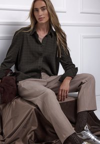 Camicia a quadri verde scuro, pantaloni beige su misura, tessuto marrone testurizzato sottostante. Scarpe da ginnastica argento con dettagli riflettenti, modello seduto in modo casuale.