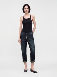 Femme mince aux cheveux courts et foncés, portant un débardeur noir, un jean foncé retroussé avec des boutons à l’avant, et des chaussures plates noires, debout sur un fond uni.