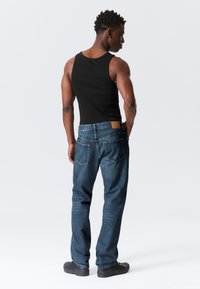 Top negru fără mâneci, cu decolteu rotund, combinat cu blugi blue denim cu o ușoară decolorare. Poză în picioare din spate, cu încălțăminte închisă la culoare.