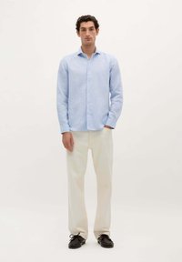 Camicia azzurra a maniche lunghe con bottone e vestibilità rilassata, abbinata a pantaloni color crema e scarpe scure. Design semplice, senza fantasie.