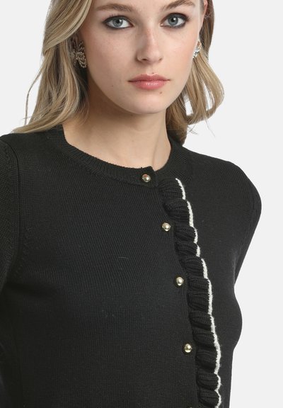 Melnā adīta kardigāna ar apaļu kakla izgriezumu, kuram ir balts viļņu akcents gar pogu rindu un zelta krāsas pogas.
